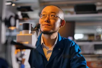 Indoor photo of Dr. Guosong Hong sitting in a laboratory, behind a microscope, looking up and to the left.