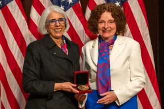 Photo of a southeast Asian woman and a caucasian woman smiling and standing together in front of American flags, holding up a gold medal in a case.