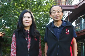 Outdoor photo of an Asian female faculty member and Asian male faculty member smiling at the camera outside of the Clark Center, wearing matching fleece vests with the Bio-X logo.