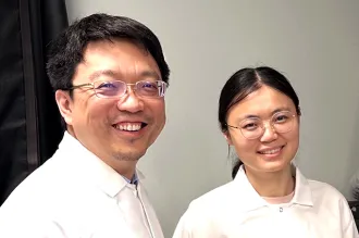 Photo of male faculty member and female postdoc standing in the lab, wearing white lab coats and smiling, with a computer screen showing cell images.