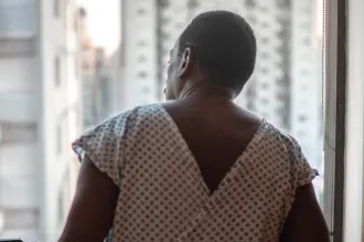 Photo of the back of a man in a hospital gown, in front of a window through which tall buildings are visible.