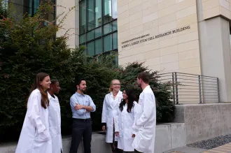 Photo of several researchers standing outside, most of them wearing white lab coats except for male faculty member standing in the middle.