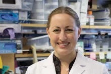 Photo of smiling female faculty member, Dr. Michelle Monje, standing in a wet laboratory space wearing a lab coat. Her blond hair is tied back.