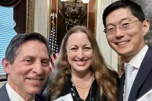 Indoor "selfie" photo of white male faculty member with short gray hair, white female faculty member with long blonde hair, and Asian male faculty member with short black hair and glasses, all smiling at the camera. They are dressed formally, and there are ornate wall decorations showing behind them.