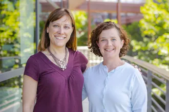 Outdoor photo of two female faculty members standing on a walkway with their arms around each other, smiling.