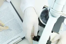 Photo from above of two scientists wearing gloves: one works with a microscope and one has a typed report on a clipboard.