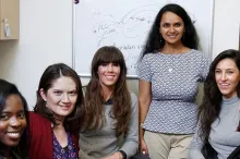 Photo of young female PI, standing, and several diverse female students from her laboratory, seated.