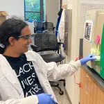 Photo of a female graduate student sitting in a wet laboratory, putting a sample intoa  machine and smiling.