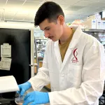 Photo of a male graduate student in a wet lab space, working with cells at a microscope, wearing a white lab coat with the Bio-X logo.