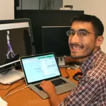 Photo of a male graduate student sitting at a computer with two screens, smiling at the camera.
