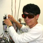 Photo of a male graduate student sitting in a wet lab space, working with electrical equipment while wearing protective goggles.