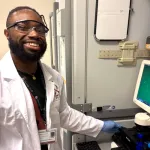 Photo of a male graduate student standing in a wet laboratory next to a monitor showing cells, wearing a white lab coat with the Bio-X logo and smiling at the camera.