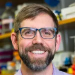 Indoor headshot photo of a smiling male faculty member with glasses, a beard, and short brown hair, Dr. Michael Jewett, Professor of Bioengineering at Stanford University.