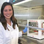 Photo of a female graduate student sitting in a wet lab by a hood, with a white box equipment inside the hood, smiling at the camera.
