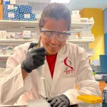 Photo of a female graduate student working at a wet lab bench with a pipette, wearing a white lab coat with the Bio-X logo and smiling.
