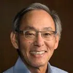 Indoor headshot photo of a smiling Asian male faculty member, Dr. Steven Chu, Professor of Physics, Molecular & Cellular Physiology, and Energy Science & Engineering at Stanford University.