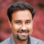 Outdoor headshot photo of a smiling Southeast Asian male faculty member, Dr. Surya Ganguli, Associate Professor of Applied Physics at Stanford University.