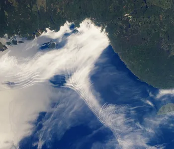 Satellite image of a large body of water and the nearby land, with numerous clouds rippling in different ways.