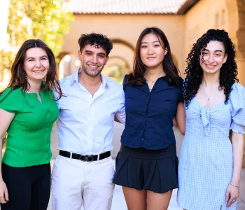 Outdoor photo of 4 undergraduate students standing together, smiling at the camera with their arms around each other.