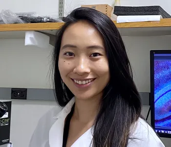 Indoor photo of a smiling Asian female graduate student standing in a wet laboratory next to a microscope, with a computer screen displaying cells in the background.