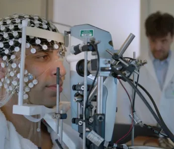 Photo of a man with a net of electrodes on his head with his face resting on a chin rest in front of a machine, with a professor in a lab coat watching from the background.