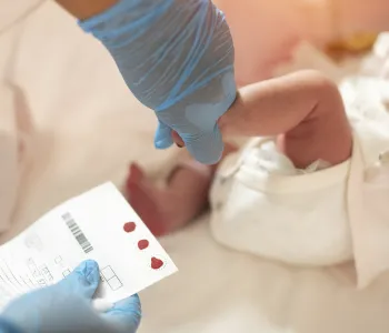 Photo of gloved hand holding a small baby's foot and other hand holding a chart with dots of blood.