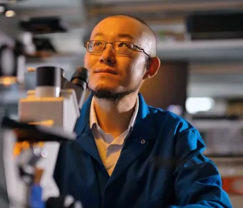 Indoor photo of Dr. Guosong Hong sitting in a laboratory, behind a microscope, looking up and to the left.