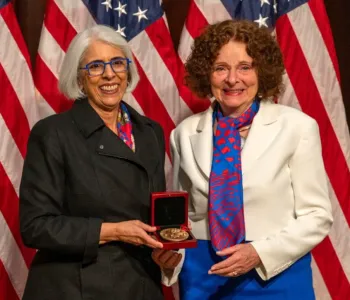 Photo of a southeast Asian woman and a caucasian woman smiling and standing together in front of American flags, holding up a gold medal in a case.