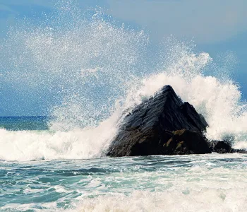 Photo of ocean waves crashing against a large rock and spraying.