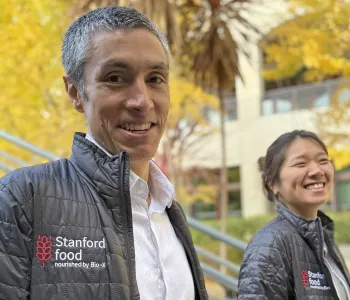 Outdoor photo of Professor KC Huang and postdoc Beverly Fu, both smiling at the camera and wearing Stanford Food branded jackets.