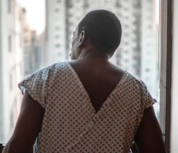 Photo of the back of a man in a hospital gown, in front of a window through which tall buildings are visible.