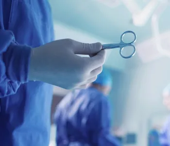 Photo of a surgeon's gloved hand holding up a pair of surgical scissors in the foreground, with operating room activity in the background.