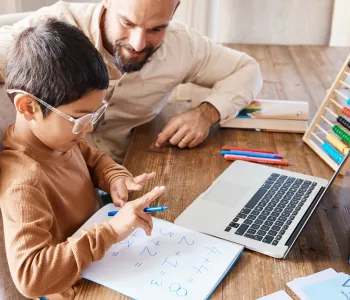 Stock photo of a man sitting with a young child who is counting with their fingers in front of a piece of paper with addition equations.