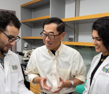 Indoor photo of Stephan Ramos, Professor Seung Kim, and Preksha Bhagchandani in the lab.