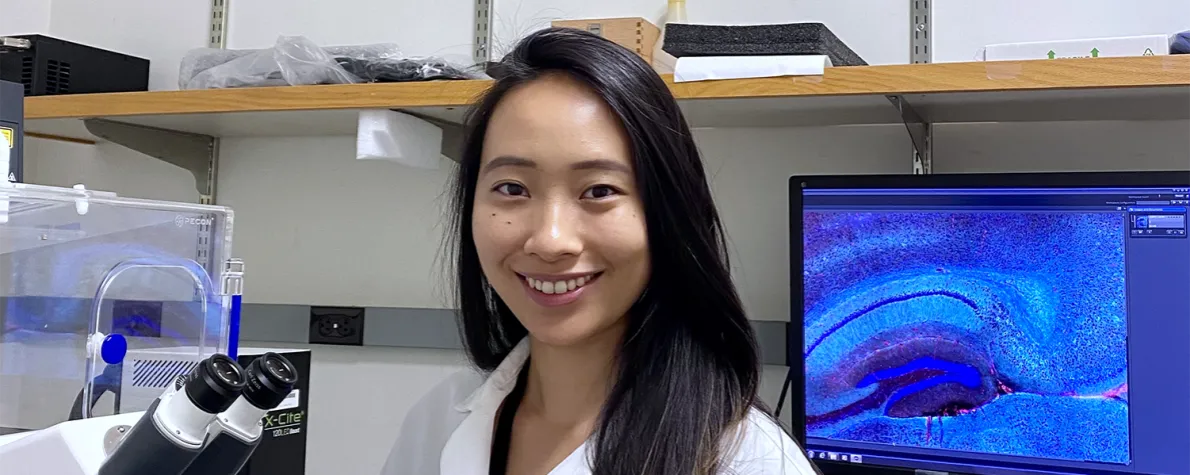 Indoor photo of a smiling Asian female graduate student standing in a wet laboratory next to a microscope, with a computer screen displaying cells in the background.