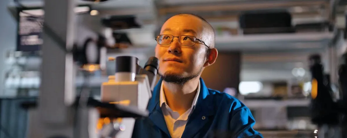 Indoor photo of Dr. Guosong Hong sitting in a laboratory, behind a microscope, looking up and to the left.