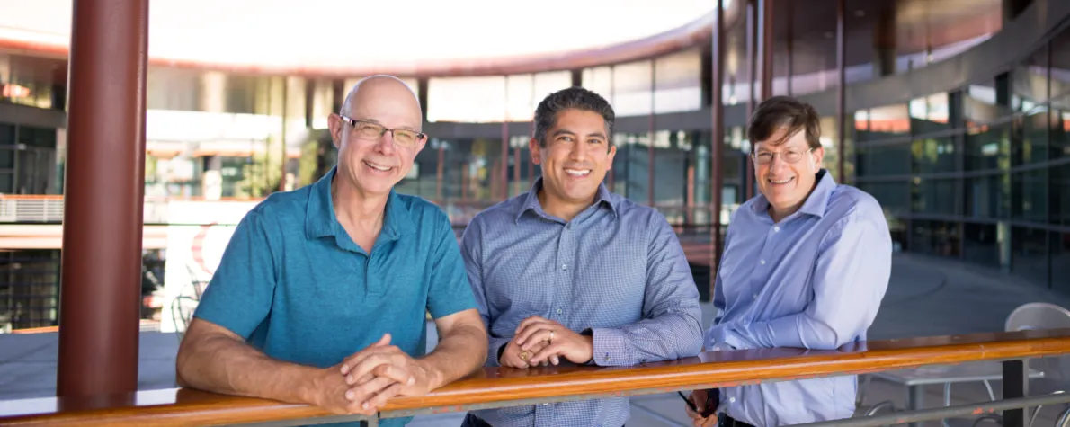 Outdoor photo of Scott Delp, Gabriel Sanchez, and Mark Schnitzer  standing at a railing on an upper level of the Clark Center at Stanford.