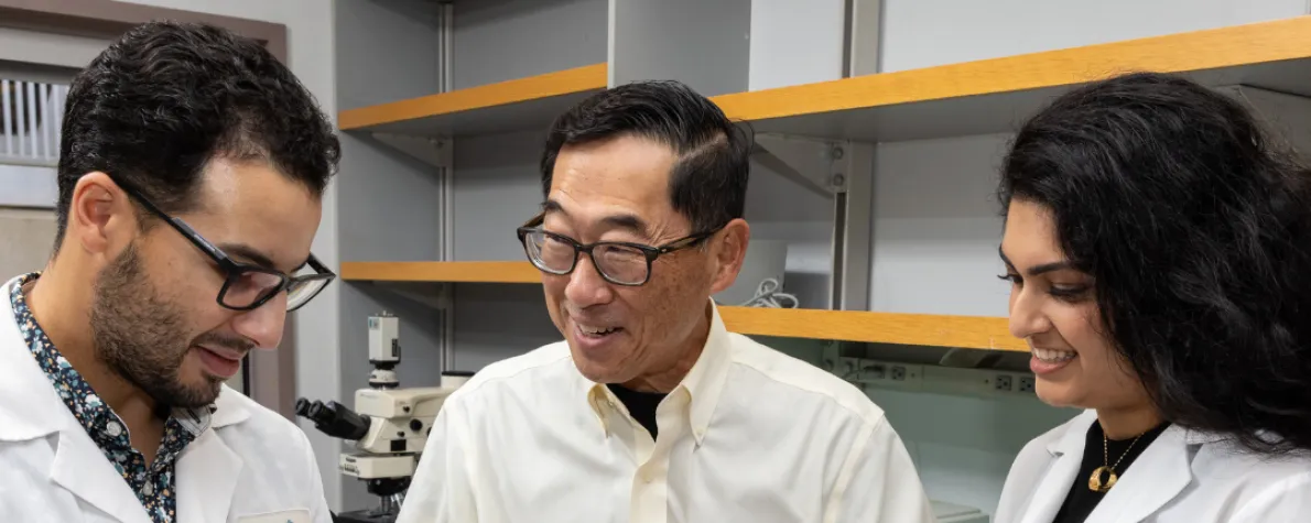 Indoor photo of Stephan Ramos, Professor Seung Kim, and Preksha Bhagchandani in the lab.
