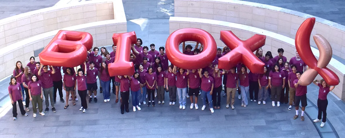 Outdoor photo of 76 students wearing matching red shirts and holding up inflated letters reading "Bio X"