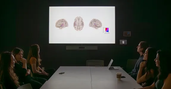 Photo of a number of people sitting around a table in a dim conference room, with brain images displayed on the projector screen.