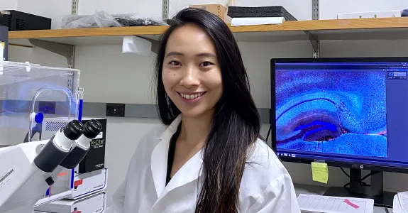 Indoor photo of a smiling Asian female graduate student standing in a wet laboratory next to a microscope, with a computer screen displaying cells in the background.