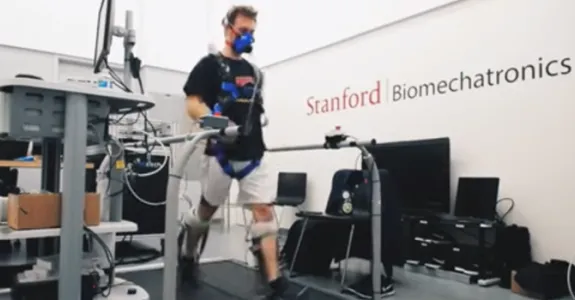 Video screenshot showing a male volunteer wearing ankle exoskeleton devices on a large mechanical treadmill, with the words "Stanford Biomechatronics Lab" on the wall behind.