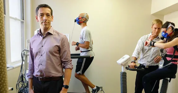 Photo of Dr. Euan Ashley standing at the front of a small room where two subjects, one male and one female, are exercising while wearing multiple kinds of sensors, monitored by a member of the Ashley lab.