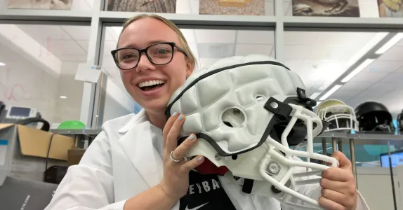 Indoor photo of a white female undergraduate student holding up a football helmet and smiling.