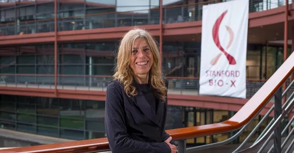 Outdoor photo of a white female faculty member standing on a staircase at the Clark Center, smiling at the camera with a large banner of the Stanford Bio-X logo in the background.