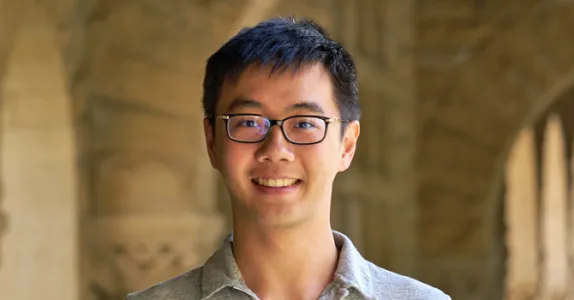 Outdoor headshot photo of a smiling male Asian graduate student with short black hair, wearing glasses.