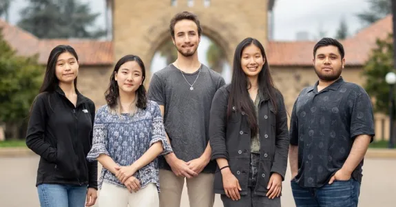 Outdoor photo of 5 undergraduates standing together in front of a Stanford University building.