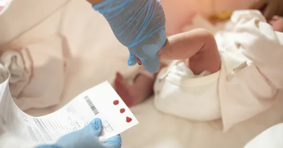 Photo of gloved hand holding a small baby's foot and other hand holding a chart with dots of blood.