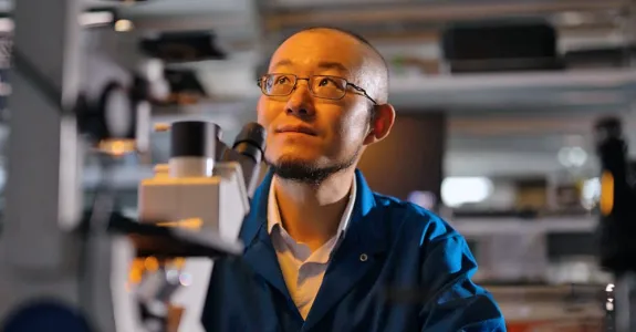 Indoor photo of Dr. Guosong Hong sitting in a laboratory, behind a microscope, looking up and to the left.