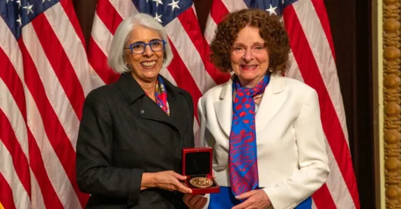 Photo of a southeast Asian woman and a caucasian woman smiling and standing together in front of American flags, holding up a gold medal in a case.